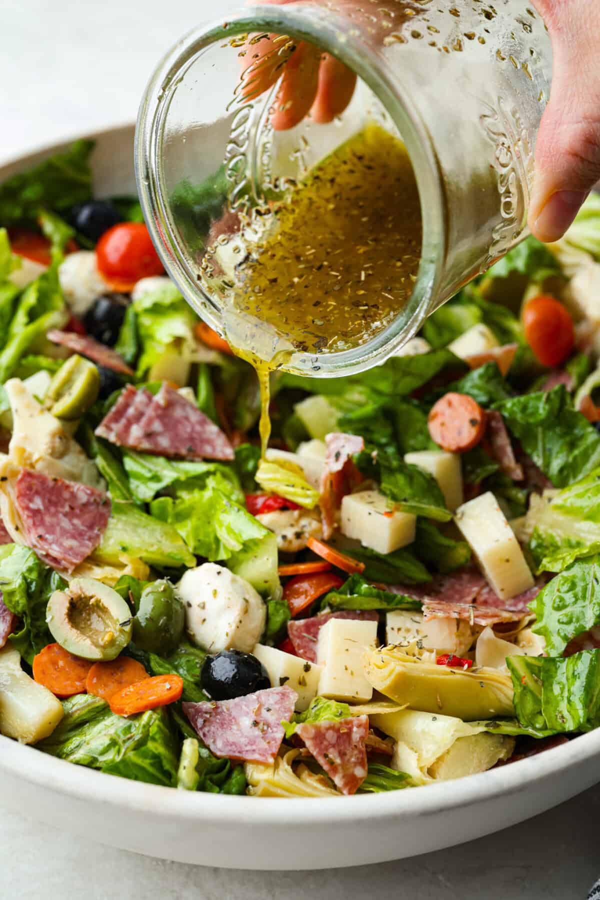 Okay, this antipasto salad? Itโs basically an Italian snack board in a bowl, and Iโmย obsessed! What makes this one a little different (and so good) is the easy lemony dressing. Someone pouring the dressing over the mixed salad.