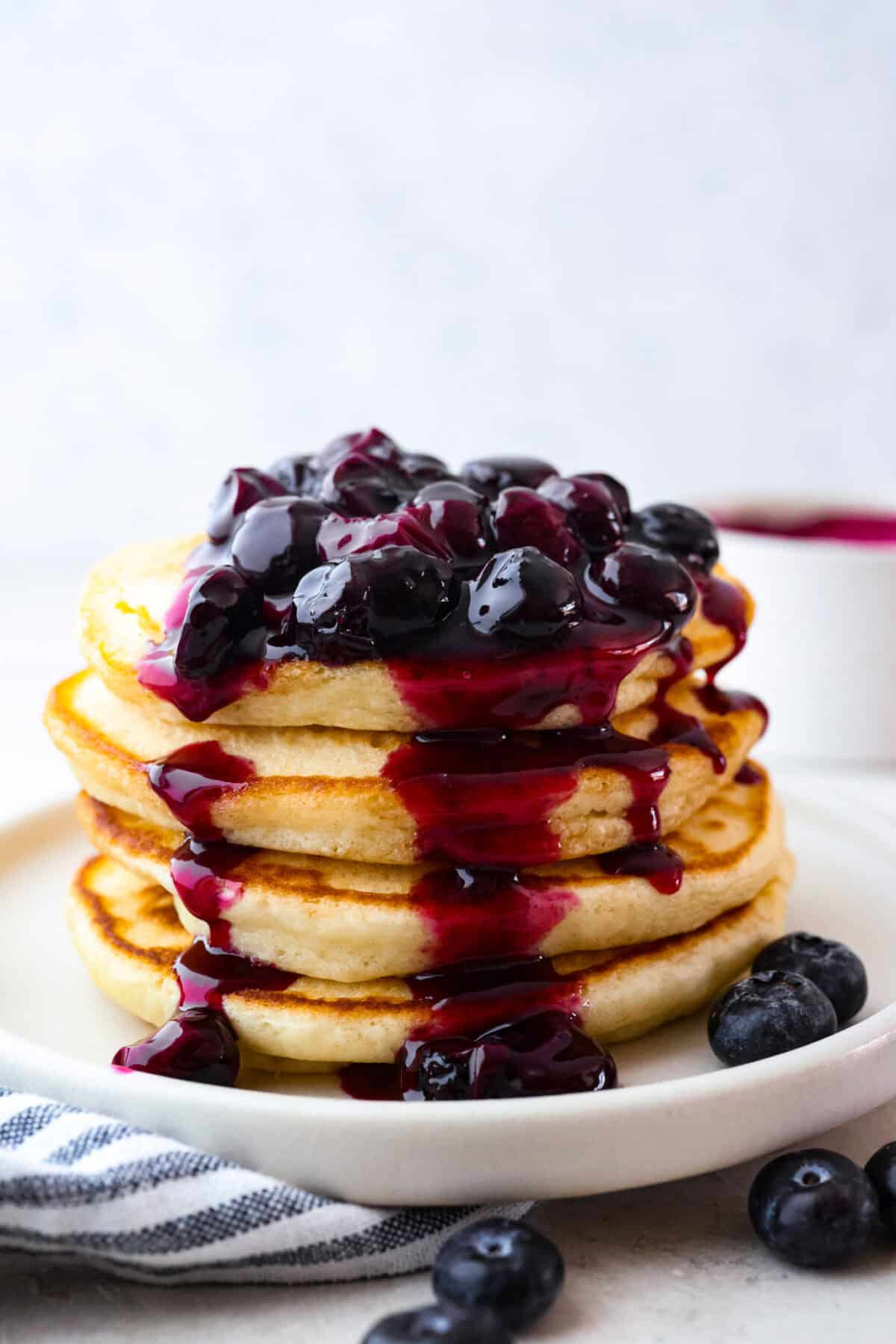 My homemade Blueberry syrup is next-level amazing! My secret ingredient? A splash of lemon! It’s the perfect balance to those sweet, juicy berries and makes your morning pancakes totally irresistible! Side shot of a stack of pancakes with blueberry syrup on top and drizzling down the sides.