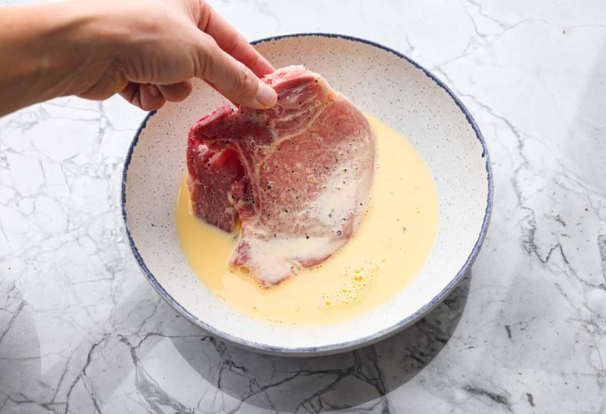 If you're looking for a tasty spin on pork chops, this is it! The crispy, cheesy texture -ย don't even get me started! YUM! Add in some roasted veggies, and you have the best Sheet Pan Crispy Pork Chops dinner! Someone dredging the seasoned pork chop into the milk and egg mixture.