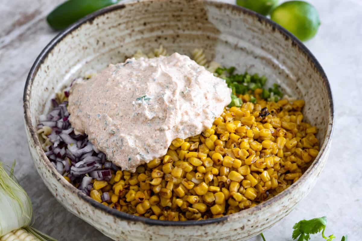 Guys, Iโm obsessed with elote, so obviously I had to turn all that smoky, creamy, cheesy, limey magic into a pasta salad big enough to feed a crowd. This oneโs guaranteed to disappear fast! Dressing added to the bowl of pasta, corn, onion and jalapeno.