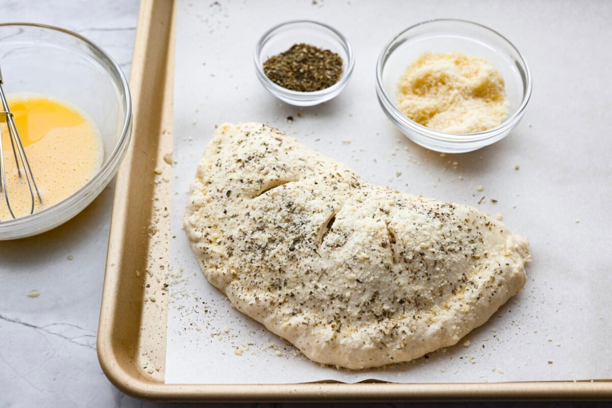 Why make calzones instead of pizza? Sometimes you just want all the pizza vibes without the mess. Calzones are like little pizza presents, warm, cheesy, and you get to make them your way! Cheese and seasonings sprinkled on top of the egg washed calzone.