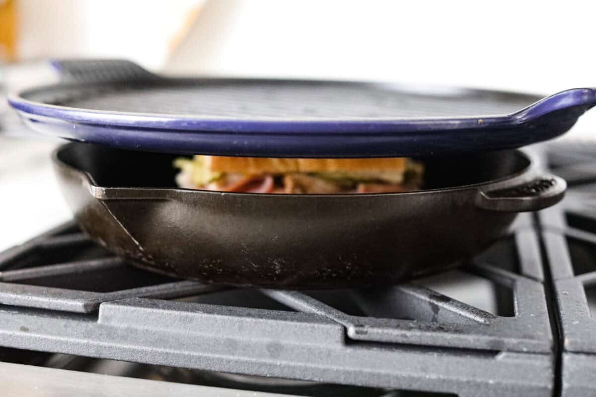 My easy Cuban sandwich is stacked with tender pork, melty Swiss, tangy pickles (my favorite), and zesty mustard! Then it's toasted in butter until perfectly crisp and golden. Heavy skillet on top of the sandwich in the skillet on the stove.