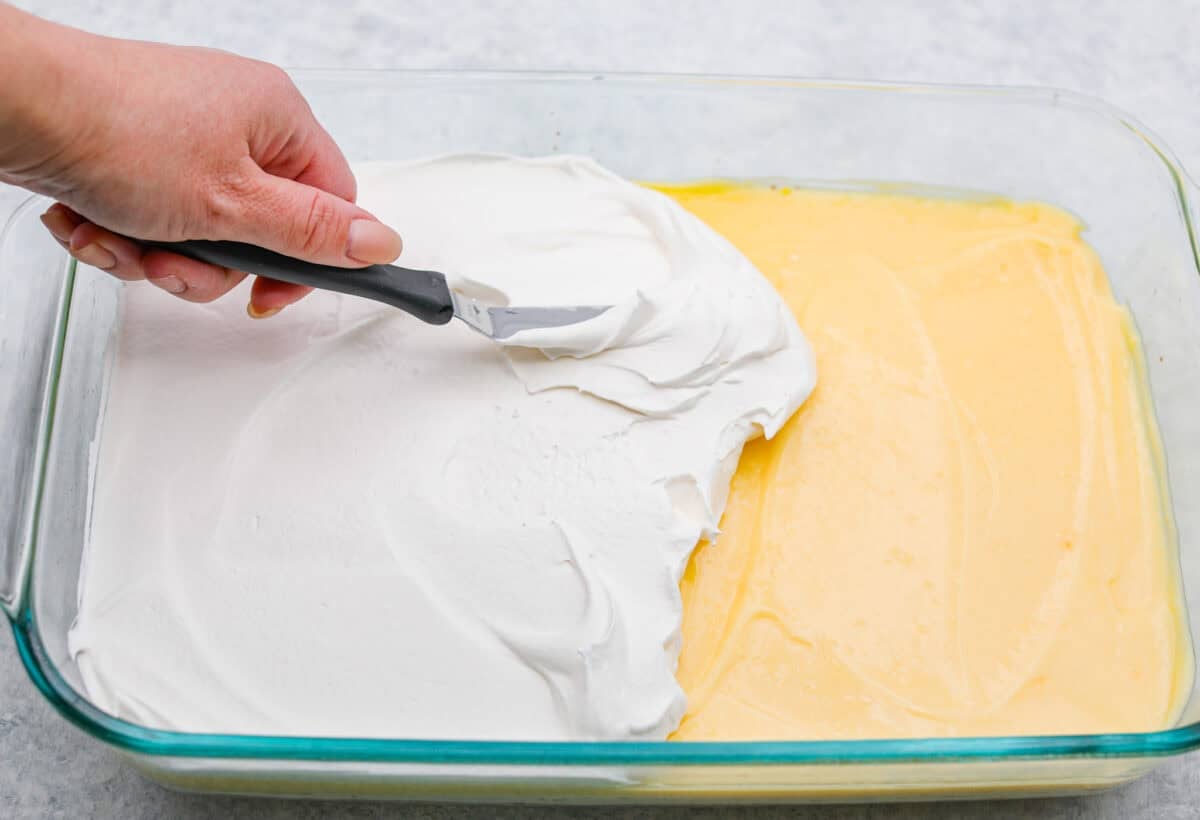 My Dad LOVES coconut cream pie! When I don't have it in me to make a pie, I make this easy Coconut Lush dessert! Itโs rich, dreamy, and always the first dessert to disappear at family gatherings. Someone spreading the whipped topping on top of the pudding layer.