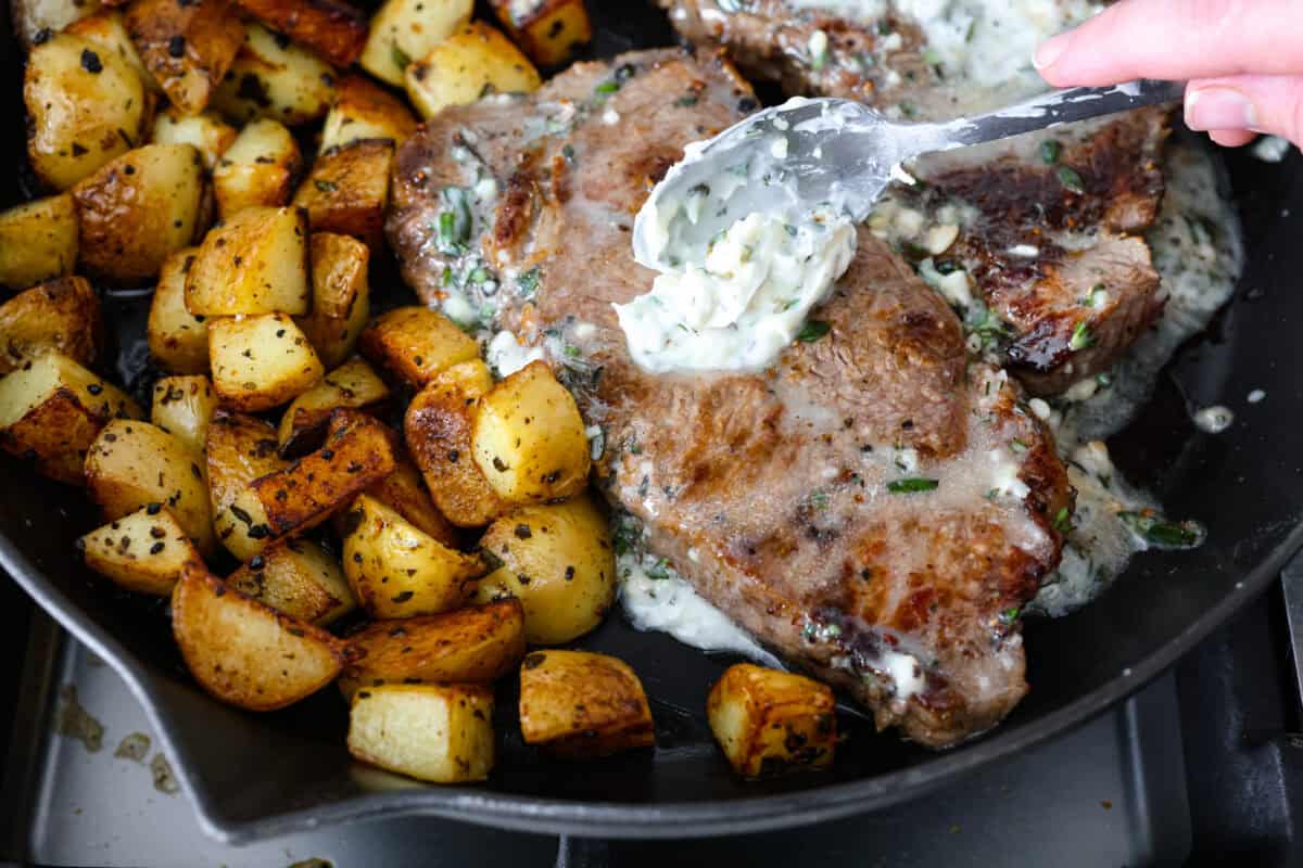If there’s one dinner that never goes out of style, it’s steak and potatoes, especially when they’re seared to perfection in a hot skillet with garlic butter and fresh herbs. This one-pan meal is full of rich, savory flavor and comes together fast. Cooked potatoes next to the cooked steaks while someone spreads the butter compound over the steak to let it melt.