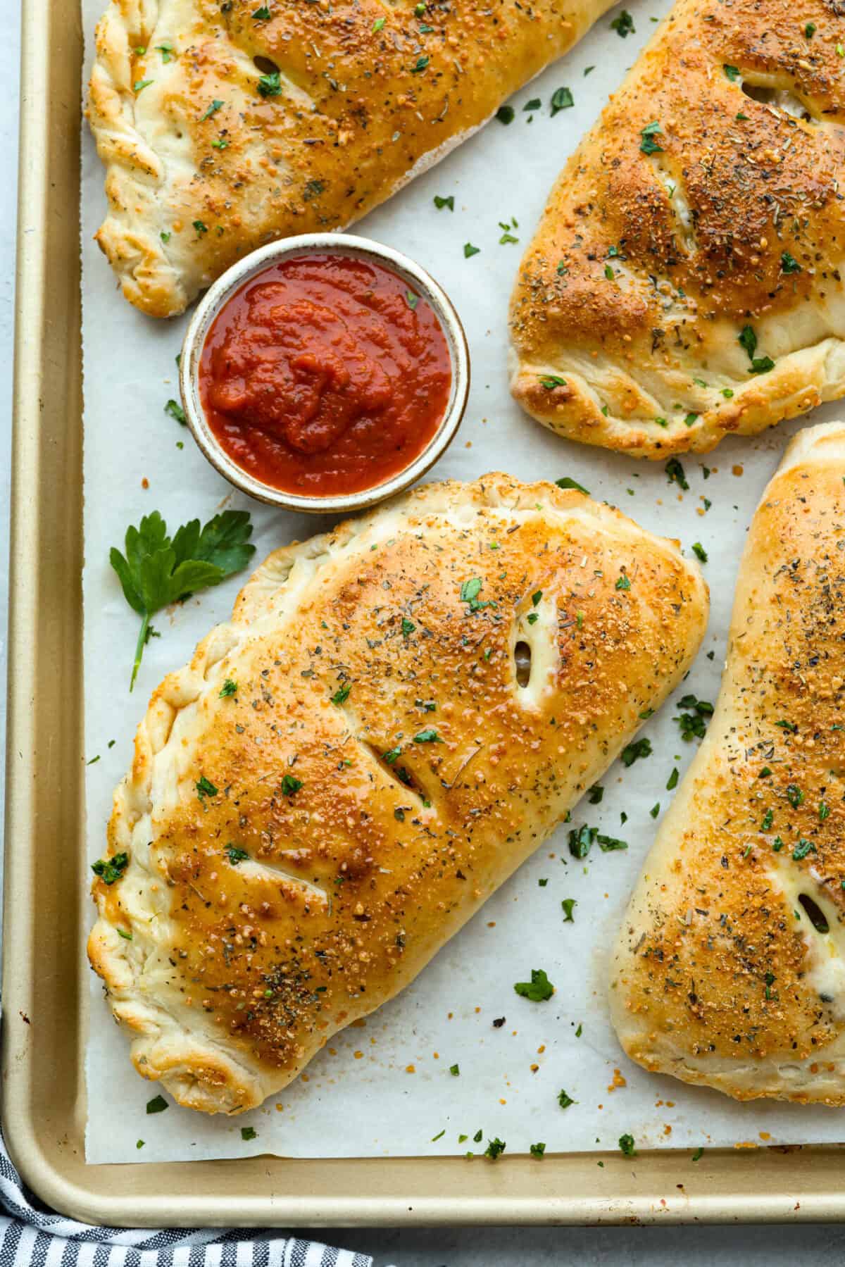 Overhead shot of beautifully baked homemade calzones on parchment paper on a baking sheet, with a small cup of marinara dipping sauce. 