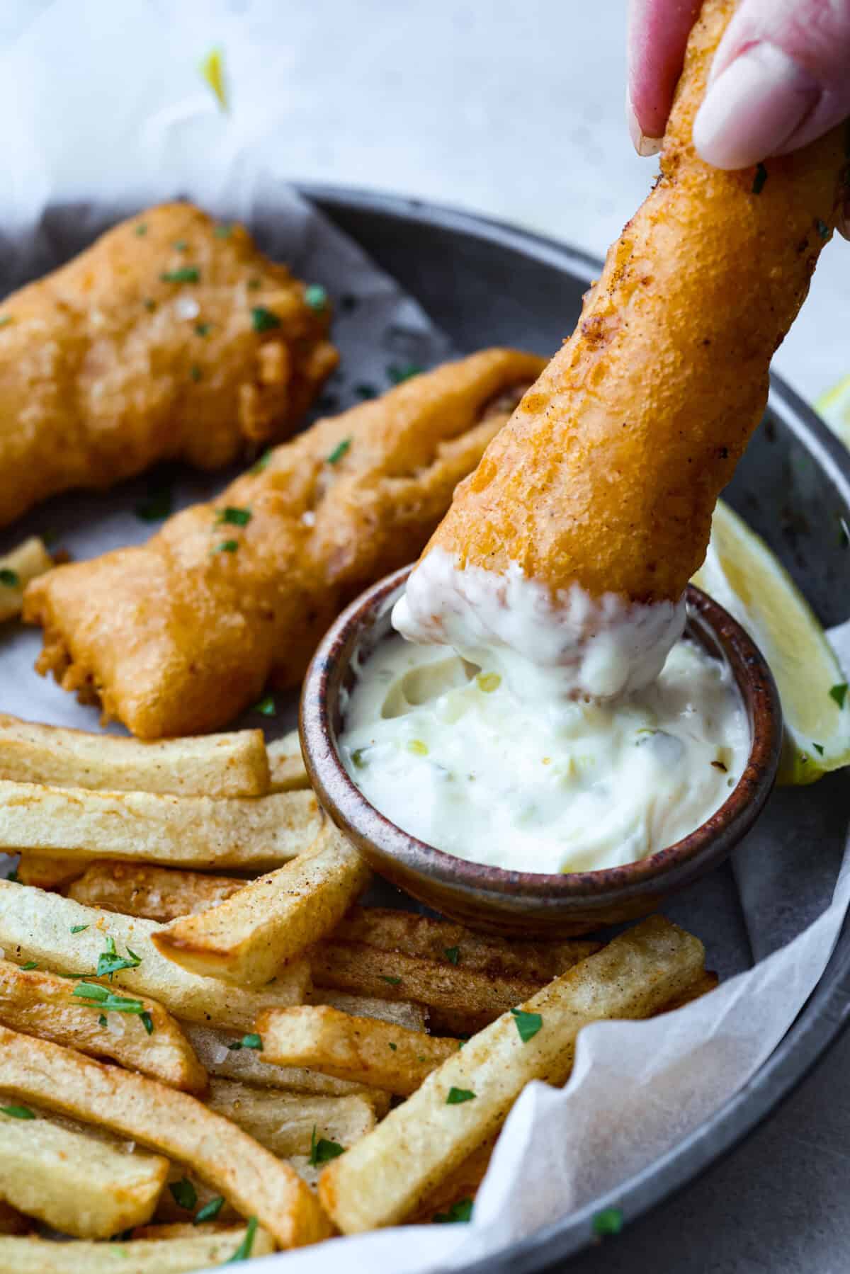 Skip the overpriced takeout and bring the pub vibes home! Crispy, golden fish and chips—light, flaky, pan-fried perfection. It’s cheaper, easier, and honestly? Way better than anything stuffed in a greasy paper bag. Someone dipping a fried fish fillet in tartar sauce.
