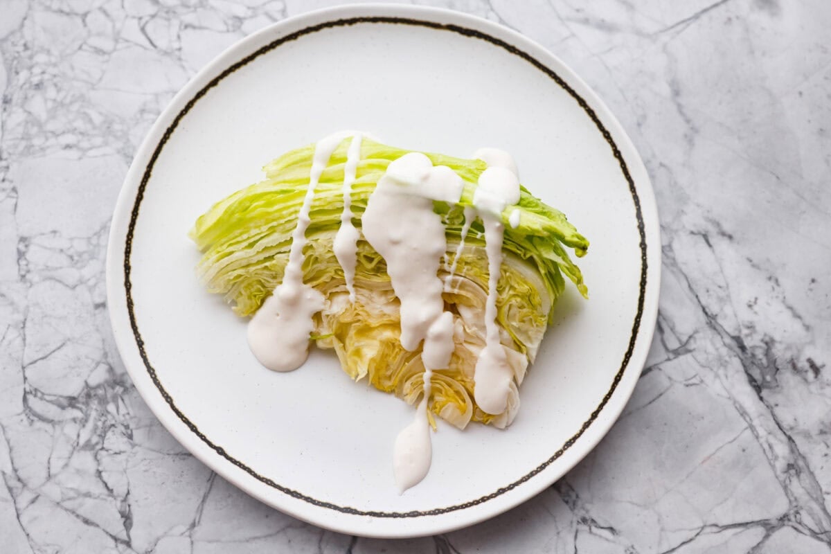 This classic wedge salad brings all the steakhouse vibes! Piled high with crispy bacon, fresh tomatoes, and a drizzle of creamy blue cheese dressing, it's a simple side that goes with just about anything. Wedge of lettuce with blue cheese dressing drizzled on top.