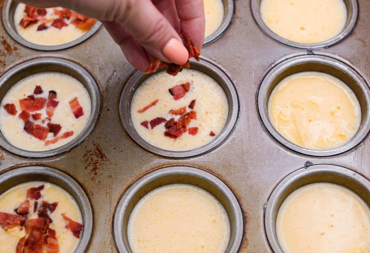 Iโve been hooked on Starbucks egg bites for years, but not the price! This homemade version is just as fluffy and flavorful, and makes the perfect grab-and-go, protein-packed breakfast. Someone sprinkling bacon over the divided egg mixture in the muffin tins.