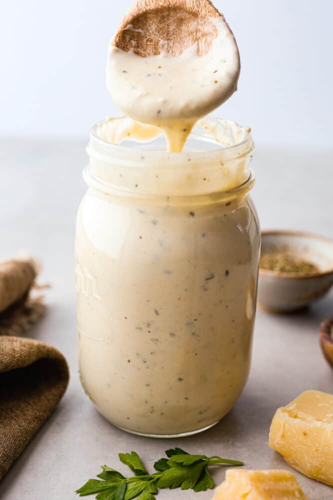 This Alfredo sauce is ultra creamy and belongs on everything. It's one of my most popular recipes EVER! It's so much better than store-bought and only takes 20 minutes to make! Jar of Alfredo sauce with a wooden spoon pouring sauce into it.