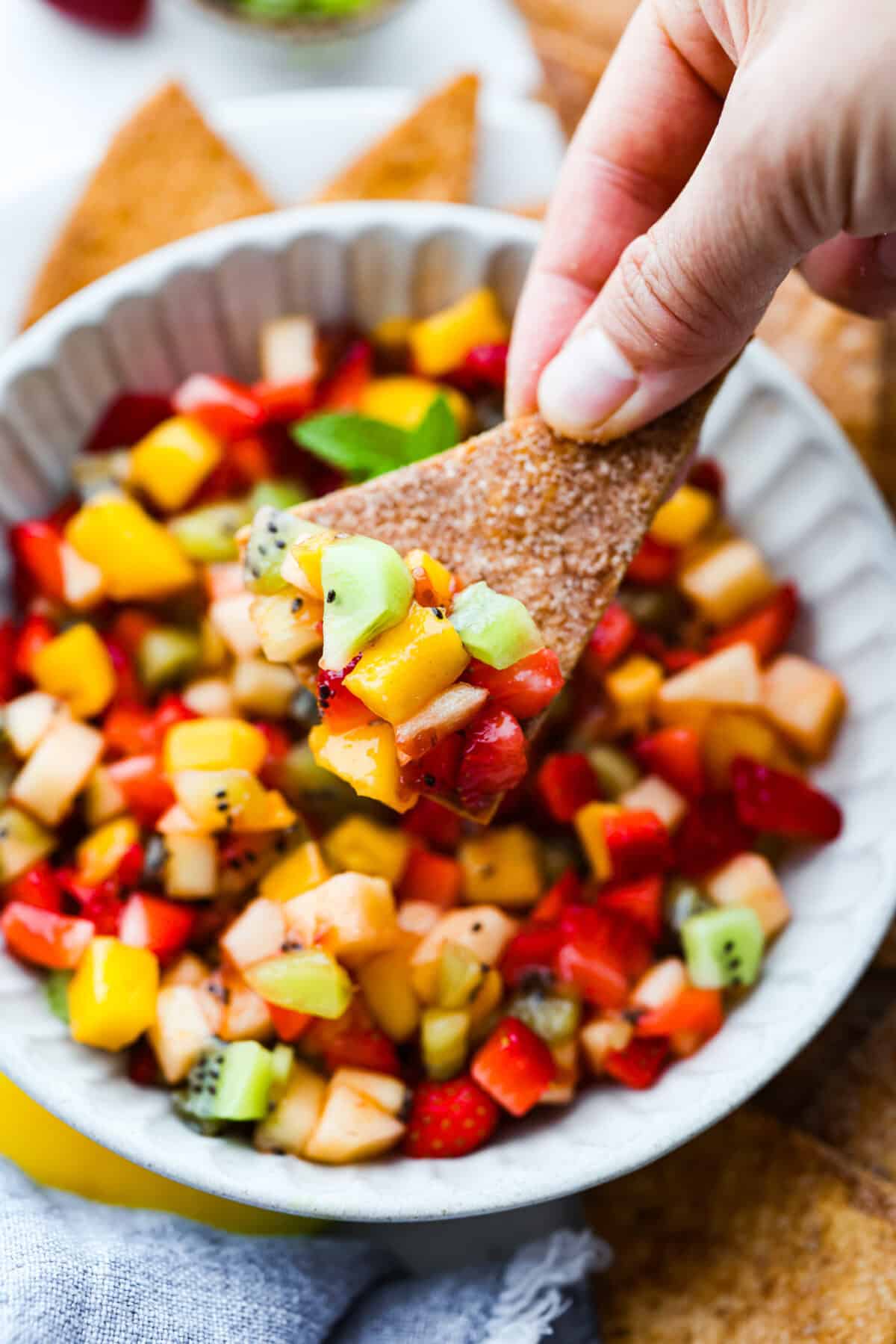 Vibrant fruit salsa is a fresh, juicy twist on your favorite snack! Sweet berries, tropical mango, and a splash of lime alwaysย steal the spotlight! Especially when served with crispyย cinnamon chips! Someone taking a scoop of fruit salsa with a cinnamon sugar chip.