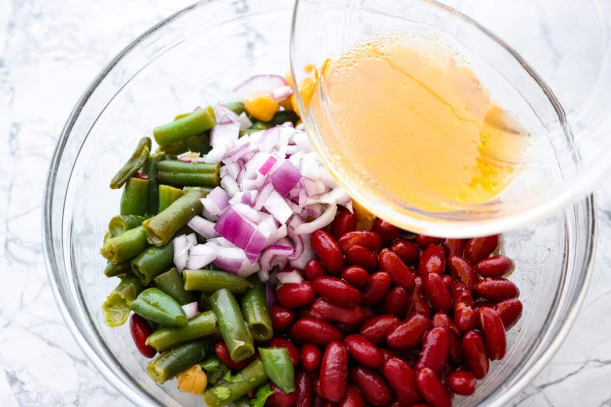 Iโve been adding fiber into my meals, and this three bean salad makes it way too easy. Itโs packed with fiber, and still tastes like a treat! It's tangy, a little sweet, and even better the next day. Someone pouring dressing over the top of the three bean salad.