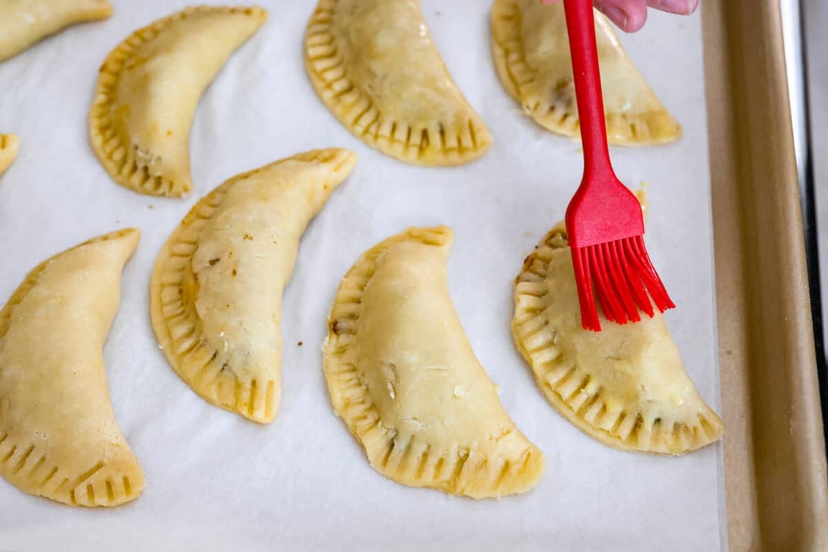 Okay, I just made homemade empanadas and Iโm obsessed! Crispy golden pockets stuffed with beef, potatoes, peas, and all the Mexican spices. You seriously need these in your life. Someone brushing an egg wash over the folded and crimped empanadas.