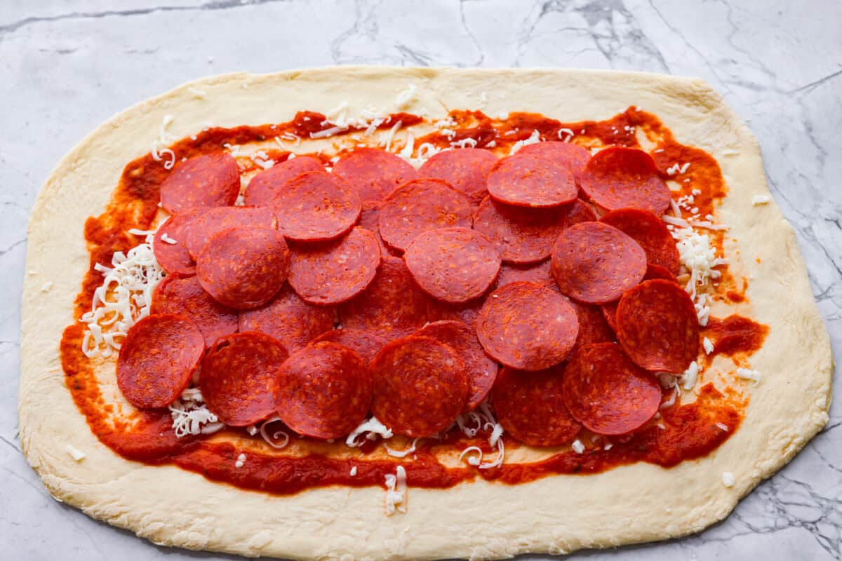 I just threw these pepperoni pizza rolls together in like 20 minutes, and my kids demolished them. AND they helped make them! You guys need to save this for a fast, fun snack or easy dinner! Sauce, cheese and pepperoni's spread out on the pizza dough.