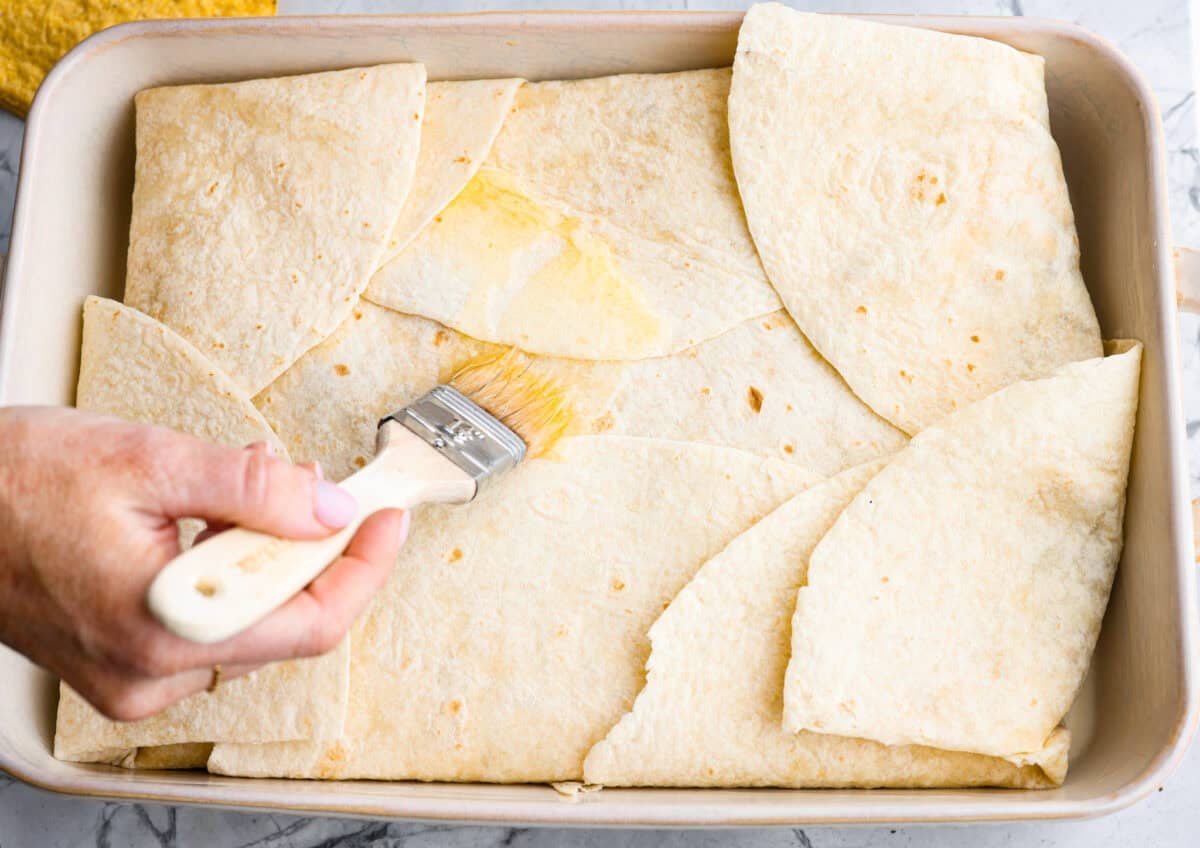 This TikTok-viral Crunchwrap Casserole takes Taco Bell’s iconic Crunchwrap Supreme and flips it into a cheesy, crunchy copycat casserole. It takes everything you love about the Crunchwrap and layers it into one big, family-sized dish! Tortillas folded in over the layers and someone is spreading butter on the the crunchwrap casserole.