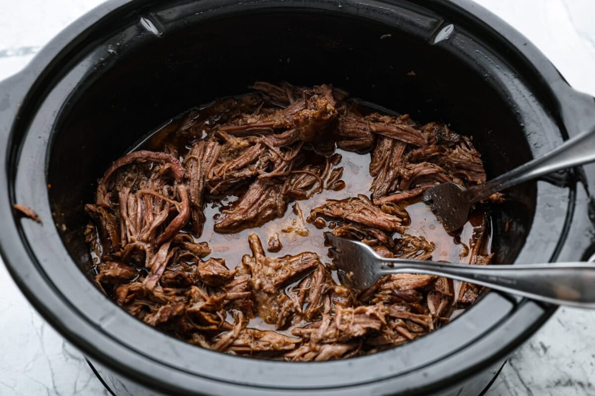 Slow cooker barbacoa beef simmers all day in smoky chipotle, tangy lime, and warm spices until it falls apart. Itโs perfect for tacos, burritos, bowls, or nachos, all with that taquerรญa-style flavor at home. Someone breaking apart the beef with forks in the slow cooker.