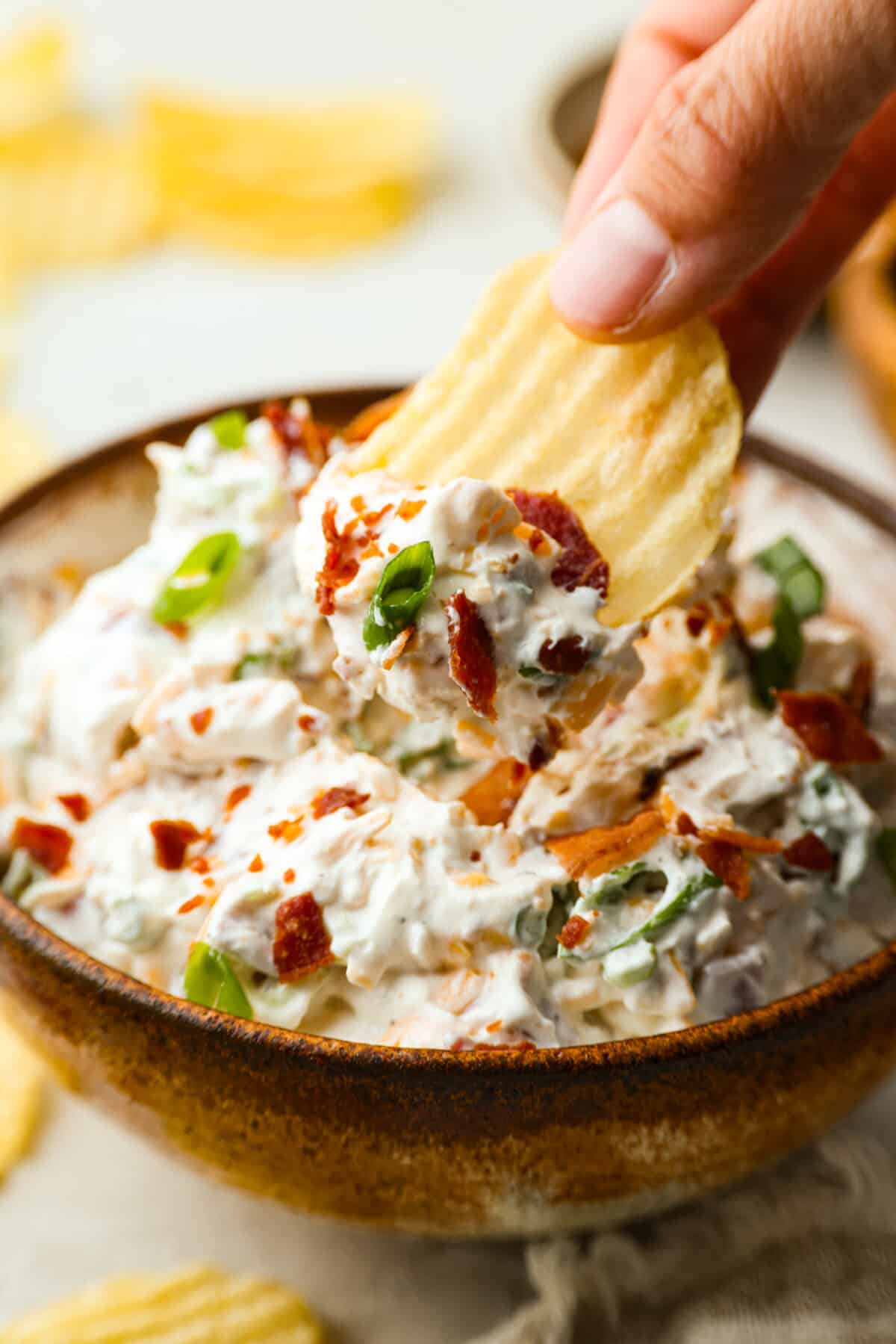You need to stop what youโre doing and make this loaded baked potato dip right now! Itโs creamy, cheesy, bacony perfection! Literally to die for! It's perfect for game day, parties, or to snack on while you catch up on your favorite show. Someone using a ruffled chip to take a scoop of loaded baked potato dip.