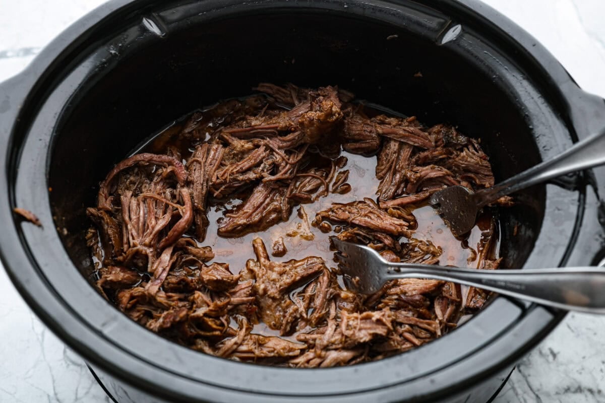 This Mississippi pot roast is melt-in-your-mouth tender, full of rich flavor, and couldnโt be easier. Just five ingredients and your slow cooker does all the work! It's a Sunday favorite at our house, and I know you'll love it too! Pot roast cooked and someone is shredding it with forks.