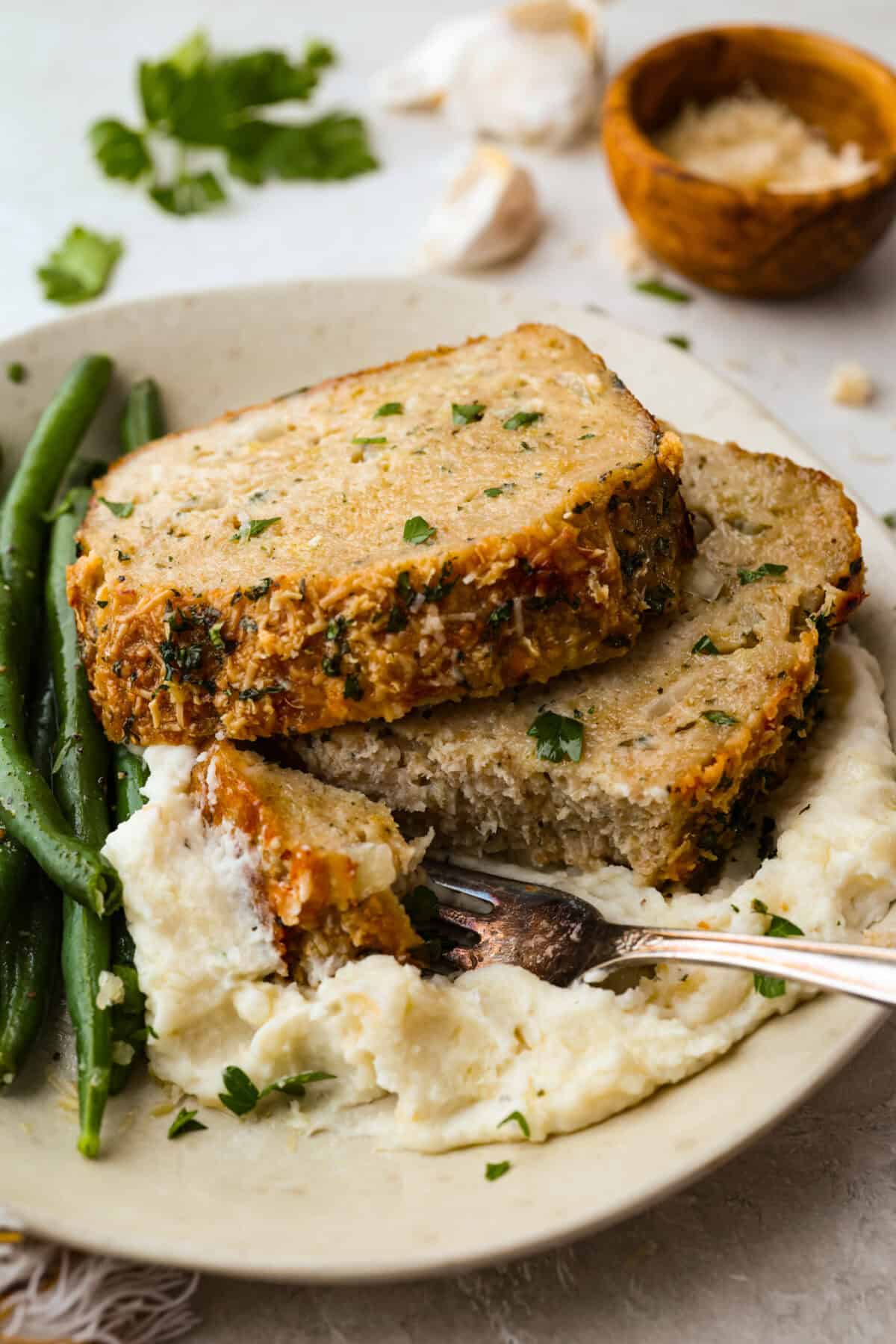 Meet your new favorite meatloaf! The ground chicken makes the Garlic Parmesan Chicken Meatloaf super moist, and you'll love the Parmesan herb-crusted topping! Plated slices of garlic parmesan chicken meatloaf with mashed potatoes and green beans.
