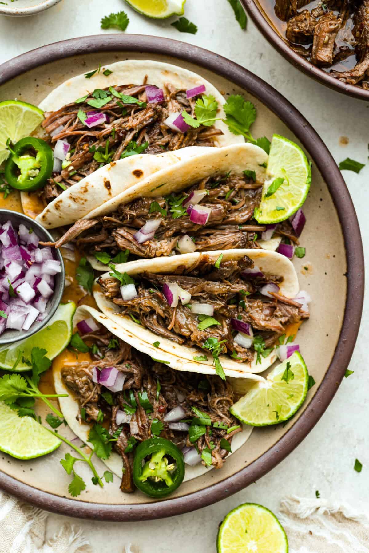 Slow cooker barbacoa beef simmers all day in smoky chipotle, tangy lime, and warm spices until it falls apart. Itโs perfect for tacos, burritos, bowls, or nachos, all with that taquerรญa-style flavor at home. Platter of tacos with slow cooker Barbacoa beef in them and garnished with red onion, cilantro, lime, and jalapeรฑos.