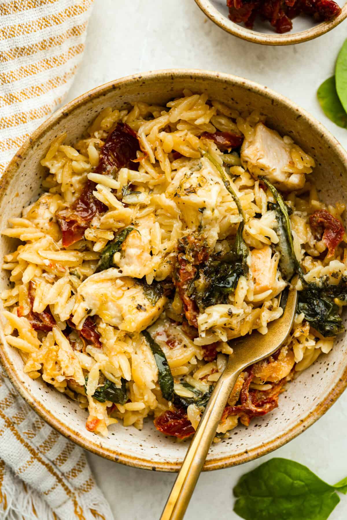 This creamy Tuscan chicken orzo bake is the kind of dinner youโll want on repeat! Tender chicken, orzo, and sun-dried tomatoes bake together in one pan for an easy, comforting weeknight meal. Bowl of tuscan chicken orzo bake with a gold fork.
