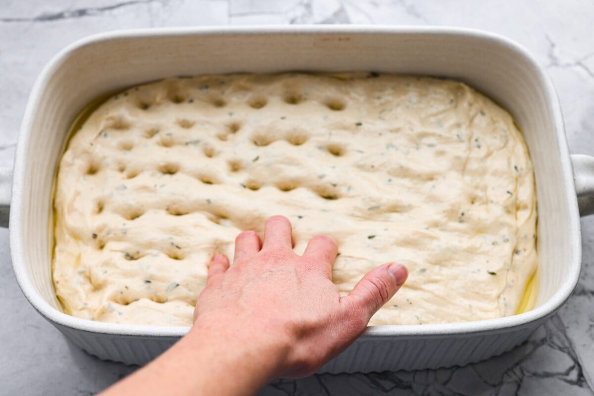 Golden, fluffy, and infused with fresh rosemary and olive oil, this rosemary focaccia is pure perfection! With its crisp edges, soft center, and irresistible herb aroma, it’s the kind of bread that disappears straight off the pan. Someone using their fingers to poke holes into the focaccia dough in a baking dish.