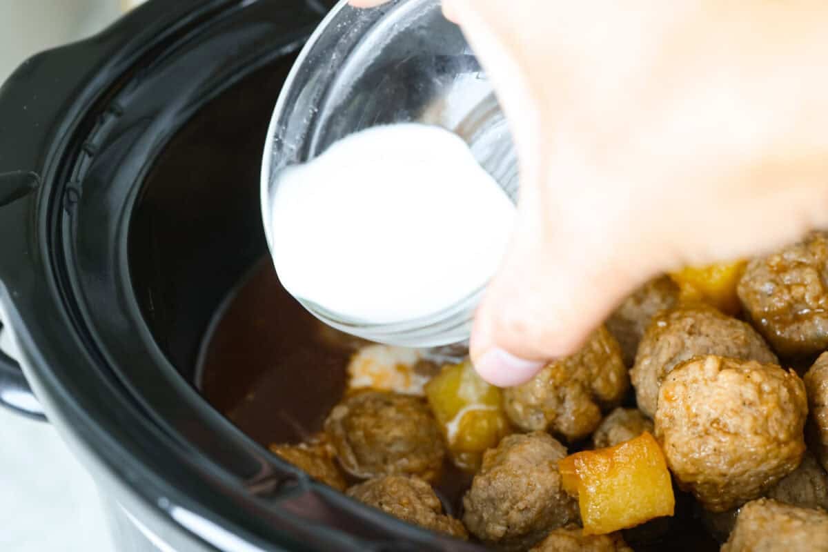 Bring the taste of the tropics to your table with Crockpot Huli Huli Meatballs! Juicy meatballs simmer in a sweet and tangy pineapple-ginger sauce, bursting with island flavor. Cornstarch slurry being added to the crockpot to thicken the sauce.
