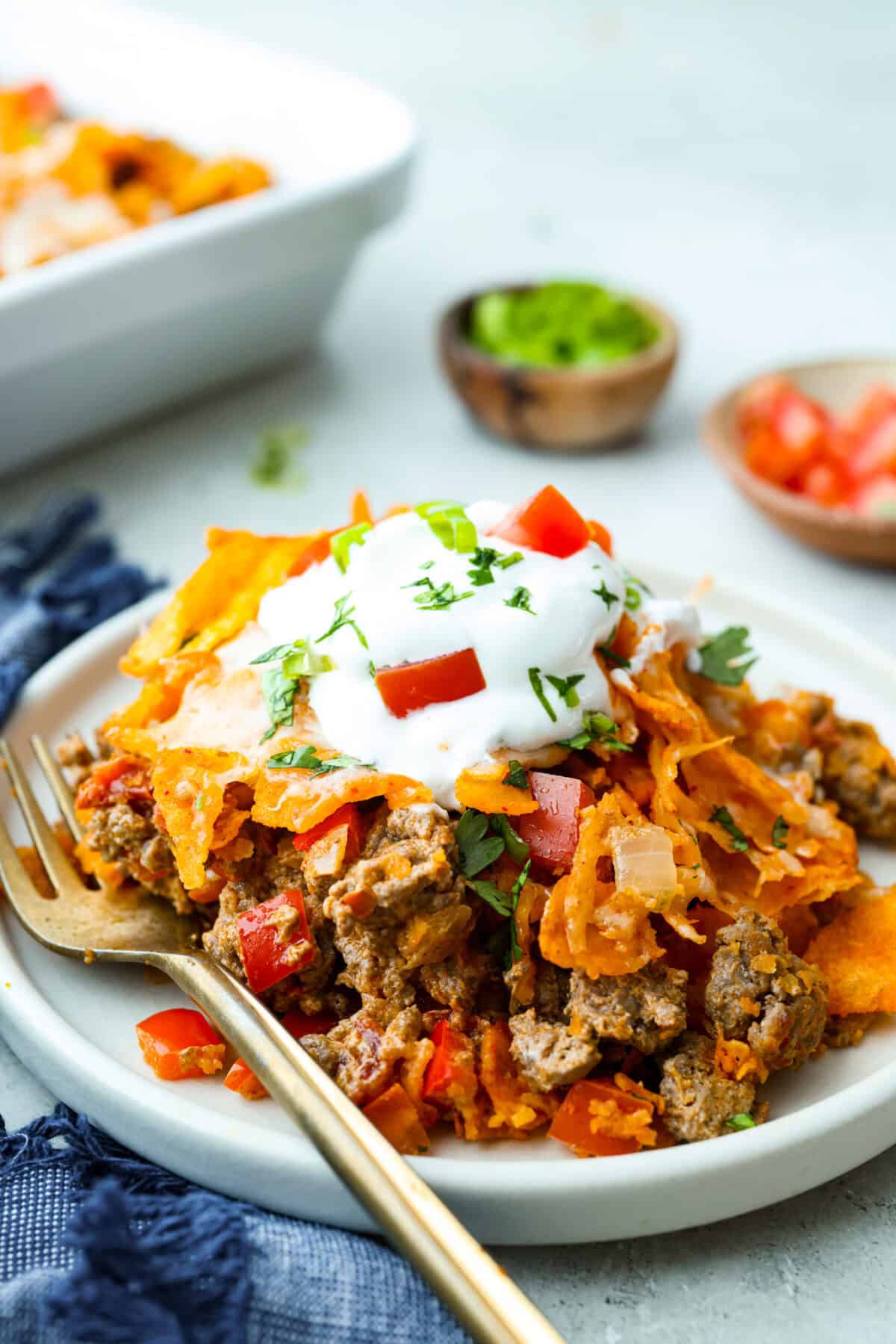 Side shot of a portion of Doritos casserole on a plate toped with sour cream, tomatoes and cilantro.