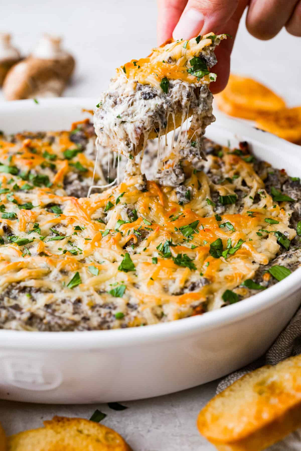 Trust me, this Stuffed Mushroom Dip is way easier (and honestly better) than making stuffed mushrooms. You skip all the filling and baking but still get that creamy, cheesy, herby, sausage flavor everyone’s obsessed with. Someone is pulling out a bite of stuffed mushroom dip.