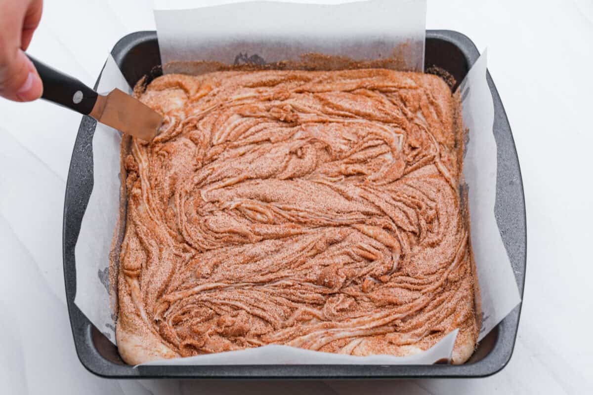 This 4-ingredient churro fudge is pure magic! It's buttery, cinnamon sugary bliss that tastes like a churro and fudge had a baby. Itโs easy to make and even easier to gift (if you can resist eating it all first!). Someone swirling the cinnamon and sugar added to the top of the fudge mixture in the pan.