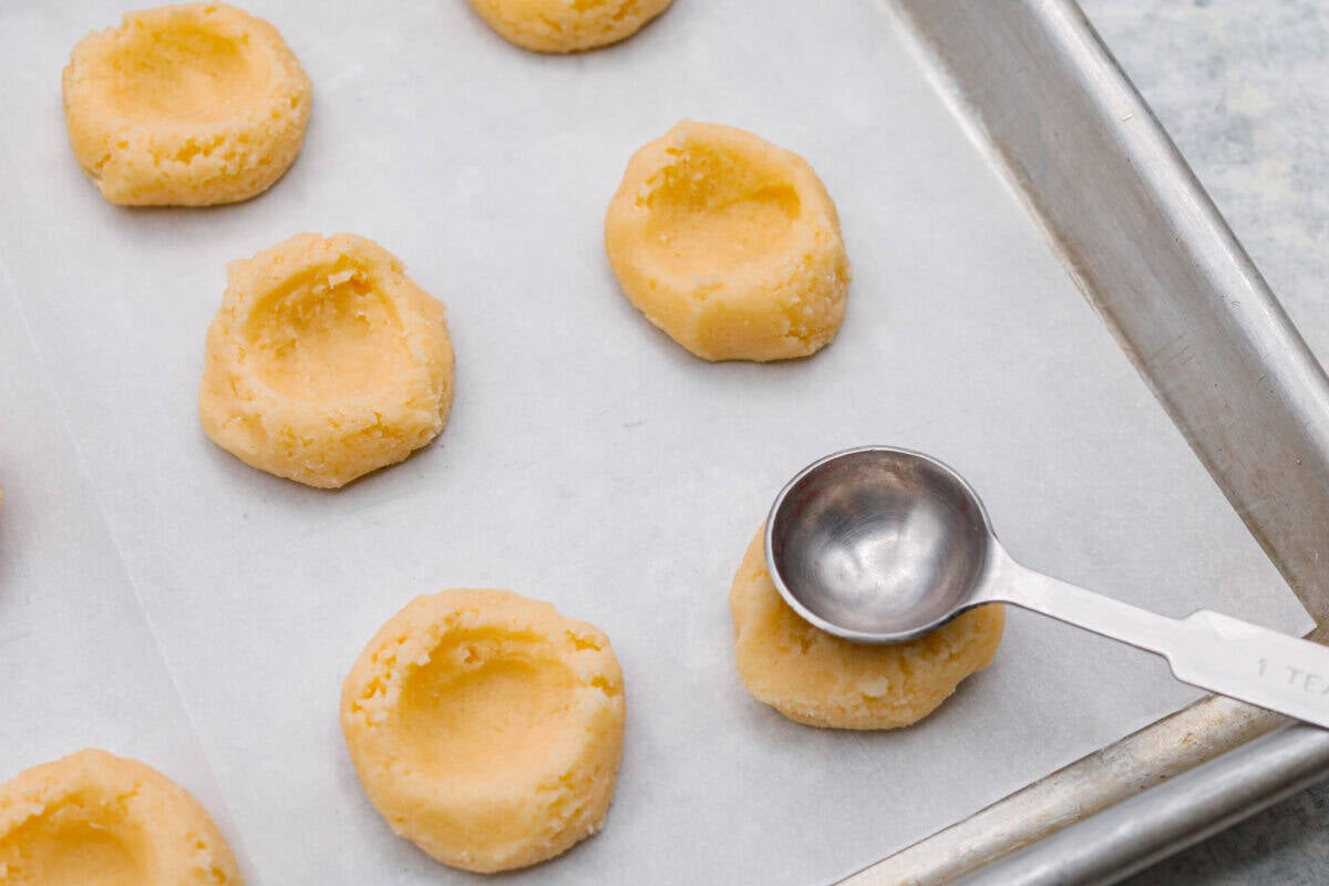Meet the upgraded Twix thumbprint cookies! I reโtested and updated the recipe so you'll get defined wells, clean edges, and a tender center every time! They are perfect for gift giving and cookie parties! Someone pressing the bottom of a rounded teaspoon into the top of the cookie dough ball before its baked.