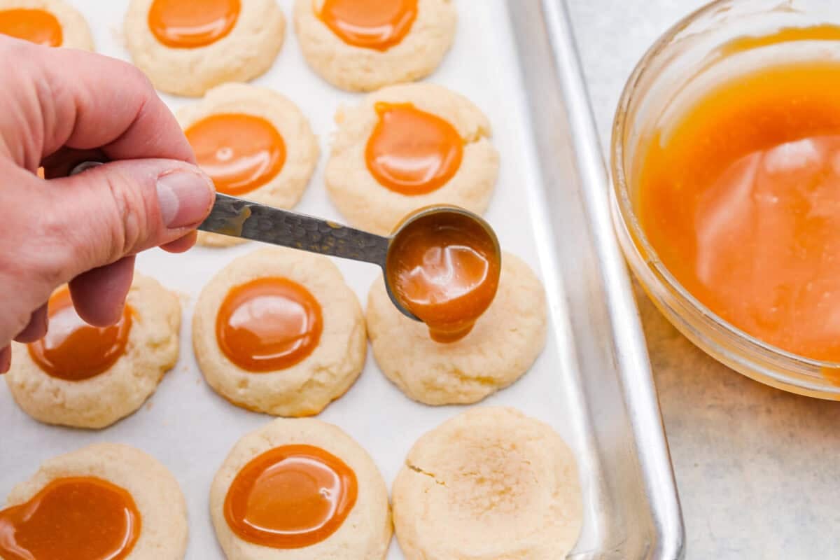 Meet the upgraded Twix thumbprint cookies! I reโtested and updated the recipe so you'll get defined wells, clean edges, and a tender center every time! They are perfect for gift giving and cookie parties! Someone spooning a small amount of caramel into the thumbprint well in the cookie.