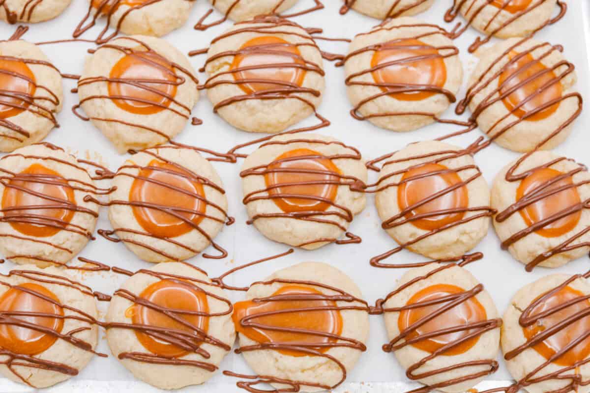 Meet the upgraded Twix thumbprint cookies! I reโtested and updated the recipe so you'll get defined wells, clean edges, and a tender center every time! They are perfect for gift giving and cookie parties! Chocolate drizzled over the top of the Twix thumbprint cookies.