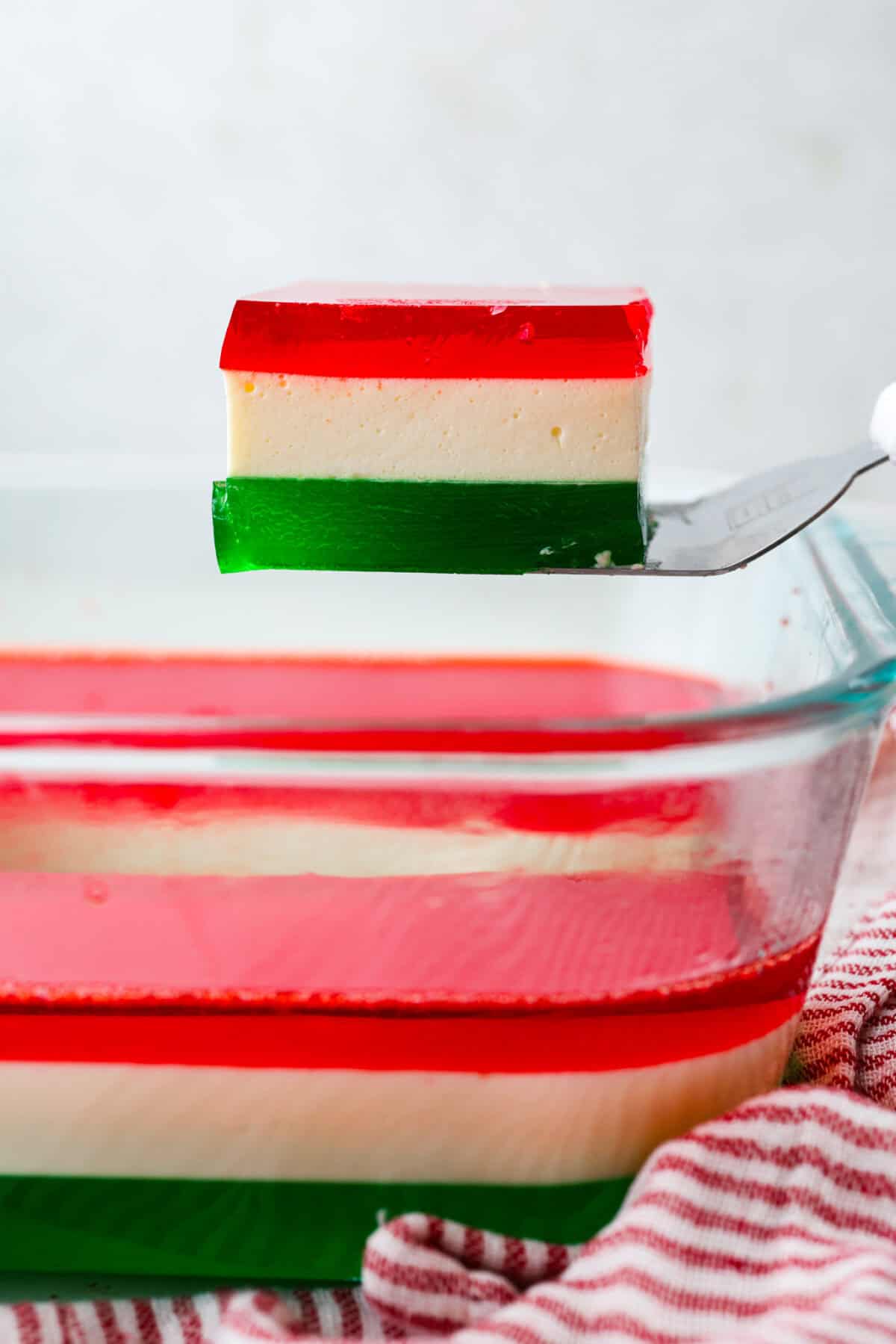 This Christmas ribbon salad brings all the festive vibes with its bright, sliceable layers. It looks fancy, but the process is super easy and mostly hands-off. Make it ahead and watch it disappear! Someone lifting a serving of Christmas ribbon salad out from the baking dish.