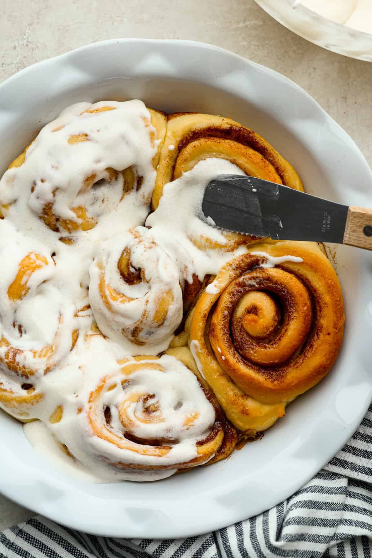 Someone spreading cinnamon roll icing over the top of baked cinnamon rolls. 