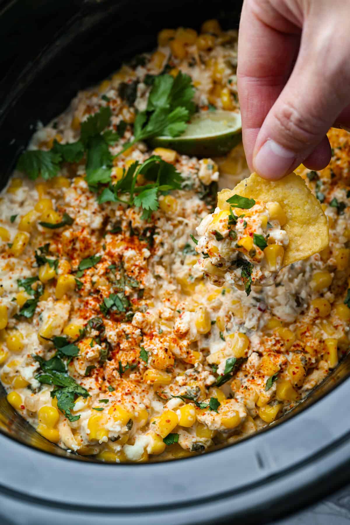 My slow cooker Mexican street corn dip takes all the flavors of elote and turns them into a warm, creamy, cheesy dip. Just toss everything in the crockpot and let it do the work. Someone scooping a portion of Mexican street corn dip with a chip.