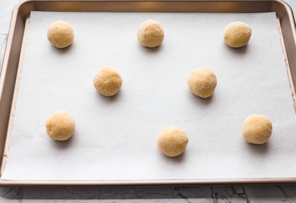 Simple to make and impossible to resist, these 4 ingredient sugar cookies are soft, tender, and sweet. A quick mix, a little roll, and your kitchen will smell like pure cookie heaven! Balls of dough rolled out and in sugar and placed on parchment paper on a cookie sheet.