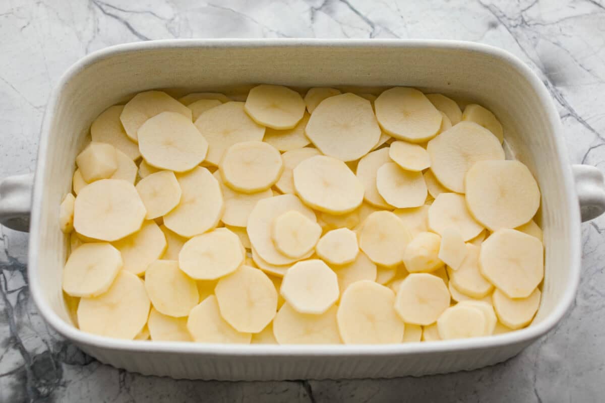 I am begging you guys to make this Zuppa Toscana Casserole! It turns the classic soup into the most delicious bake! It has layers of potatoes, sausage, bacon, kale, and cheese drenched in the most amazing sauce! Make it today, trust me! Sliced potatoes layered on the bottom of the baking dish.