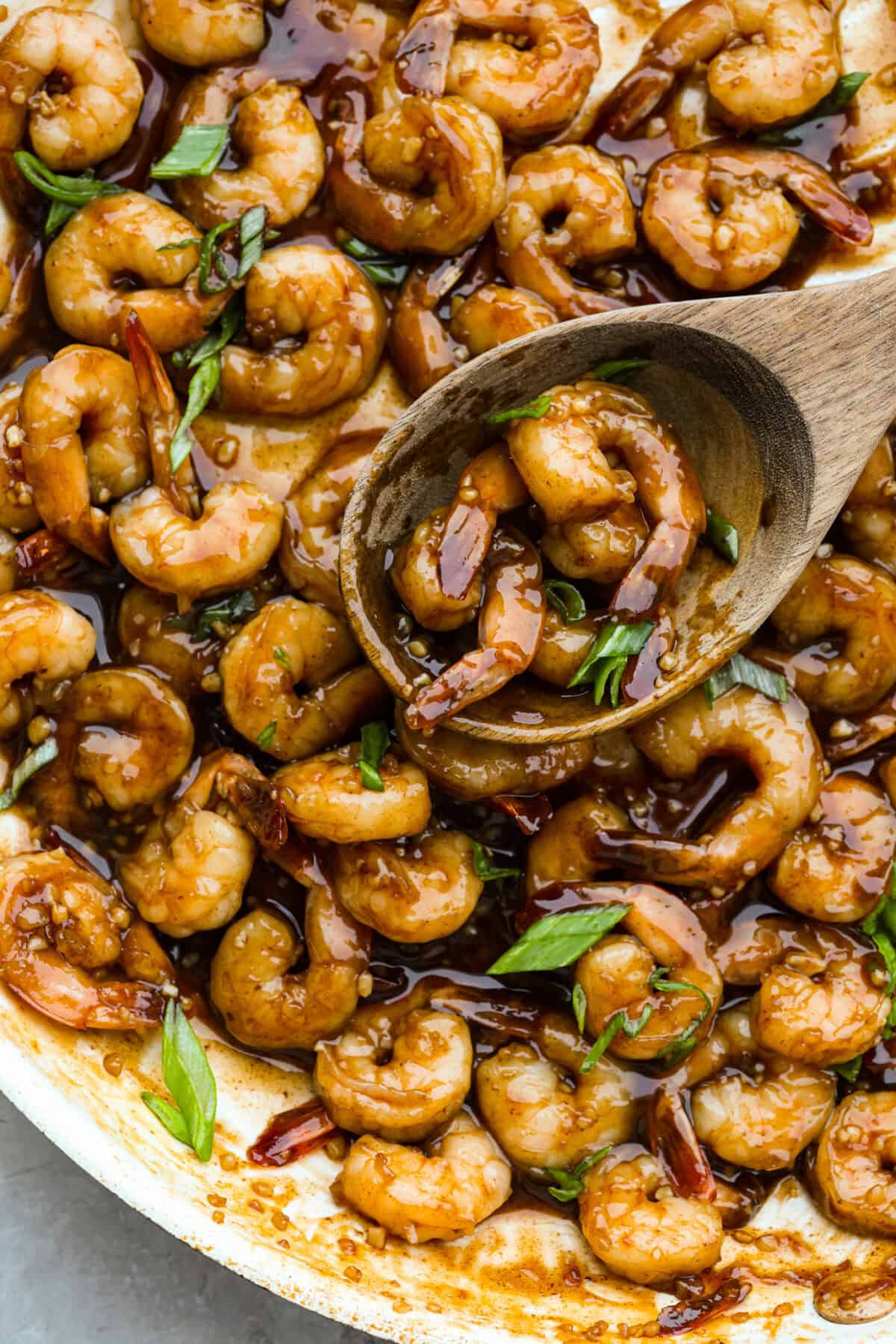 Bourbon shrimp are sticky, garlicky, and packed with flavor. Sweet brown sugar and a splash of bourbon create a quick, glossy glaze that makes this appetizer (or weeknight dinner!) totally irresistible. Someone using a wooden spoon to take a scoop of bourbon shrimp out of the skillet.