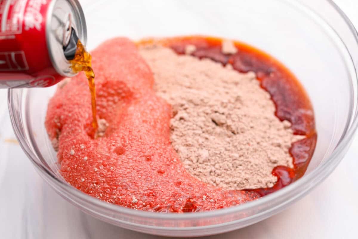 This 2-ingredient red velvet cake is almost too easy to believe! With just a cake mix and a can of soda, you can make a soft, fluffy cake in minutes. It’s the perfect shortcut dessert for Valentine’s Day or any sweet craving! Someone pouring a can of soda into a mixing bowl with cake mix.