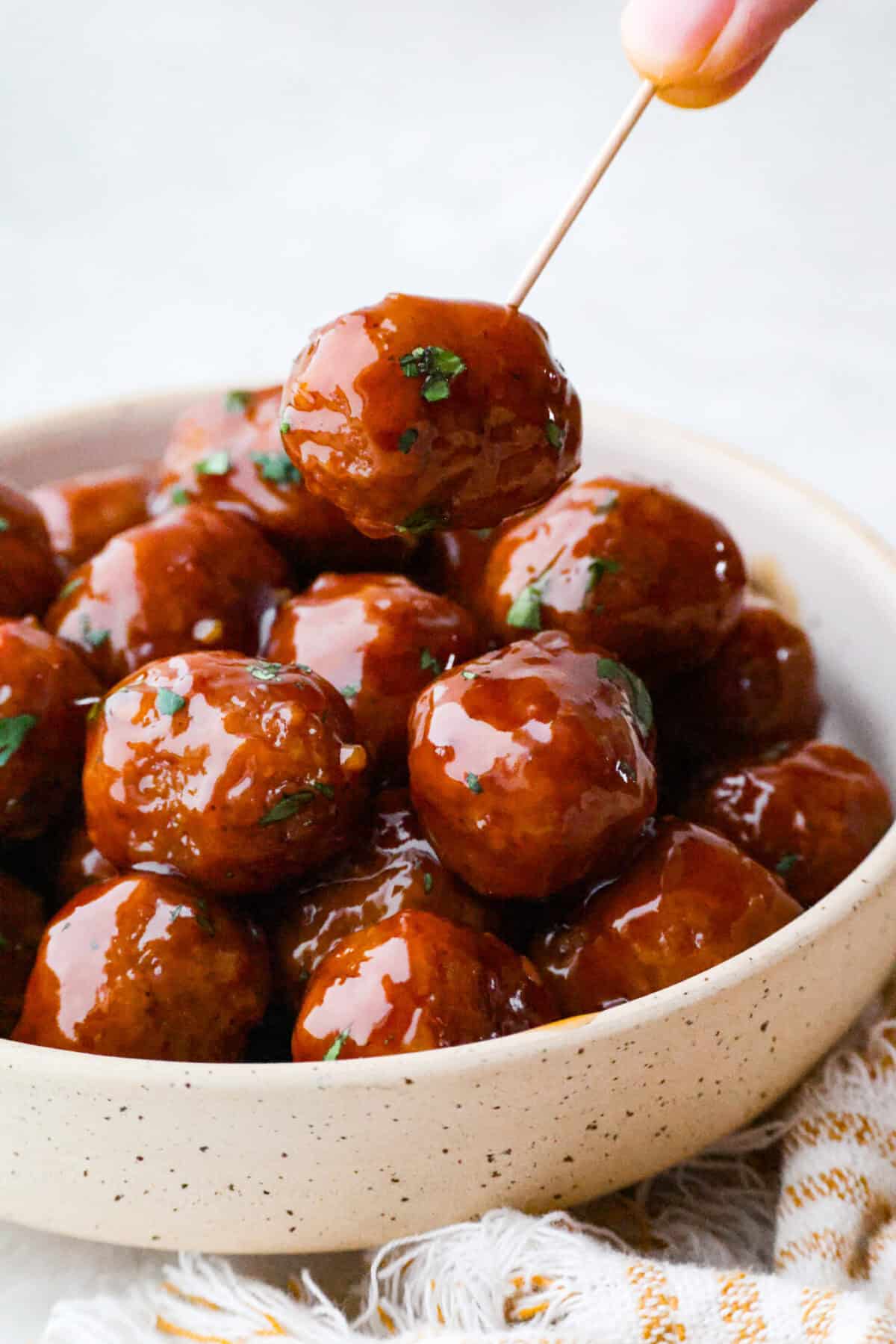 Slow cooker bourbon meatballs are an easy, set-it-and-forget-it favorite. Frozen meatballs simmer in a sweet, sticky bourbon sauce, making them perfect for an appetizer or a simple dinner with just 10 minutes of prep. Someone using a toothpick to pick up a slow cooker bourbon meatball out of a bowl.