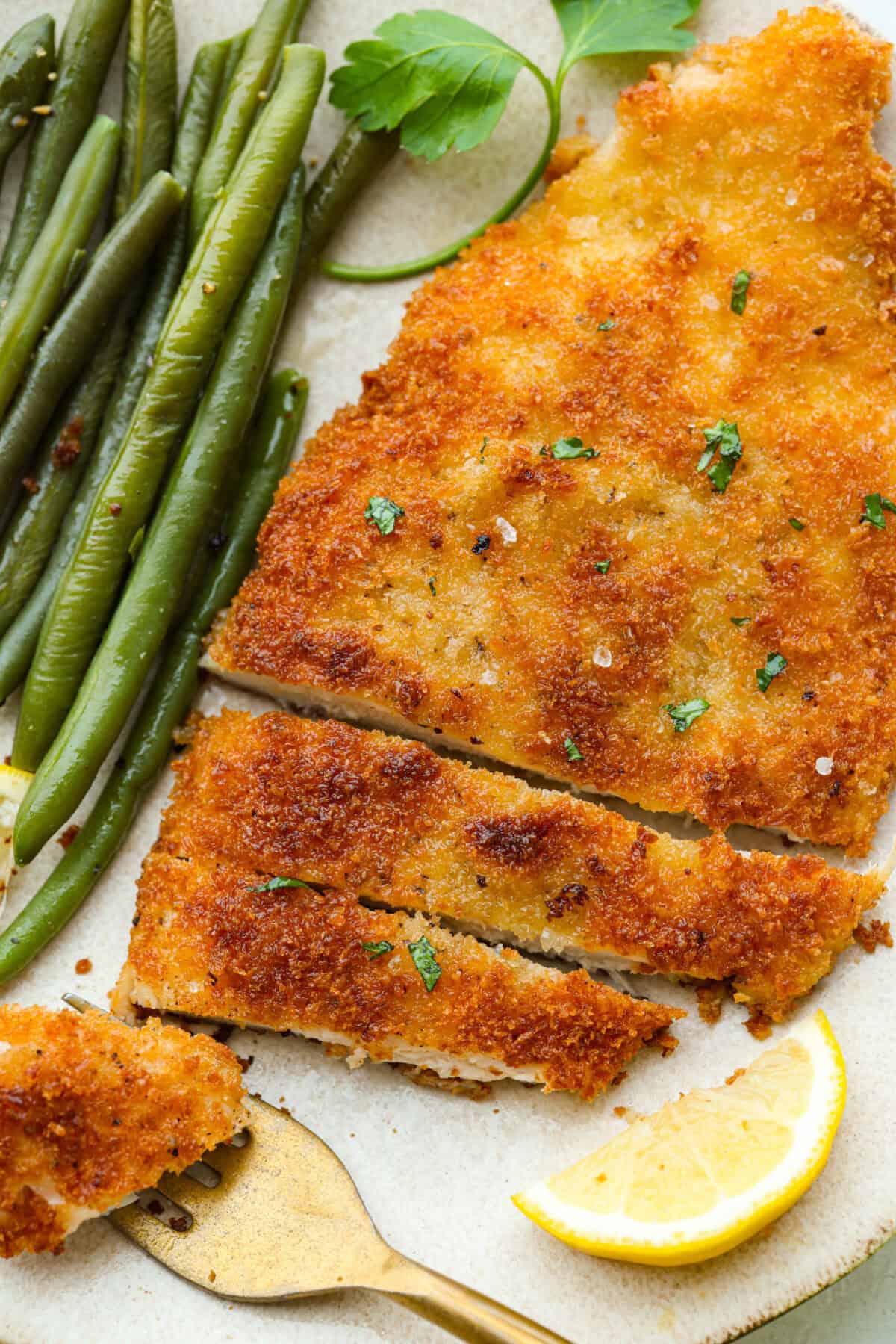 Chicken schnitzel cooked and plated next to some green beans and lemon wedge. 