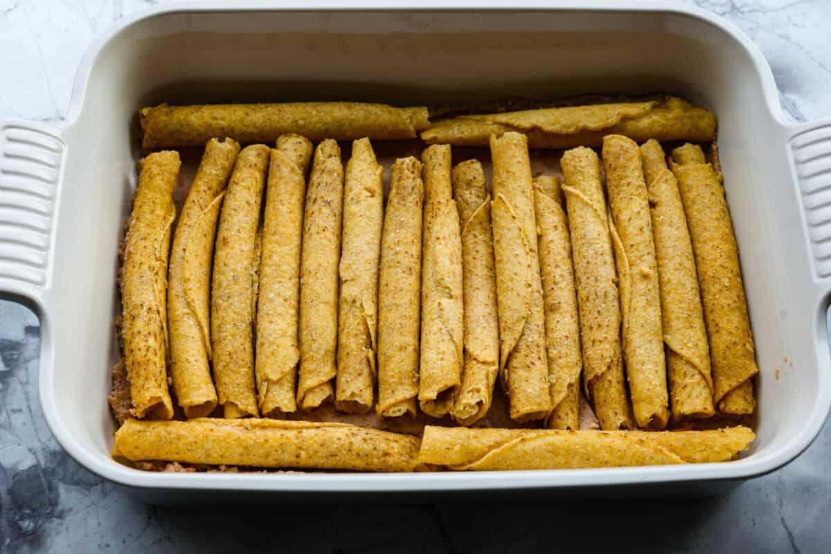 When you want enchiladas without the work, these Lazy Enchiladas are it! Four ingredients. Less than 30 minutes. Just layer frozen taquitos with beans, sauce, and cheese, bake until bubbly, and boom, dinner is served! Taquitos layered on top of the refried beans in the baking dish.
