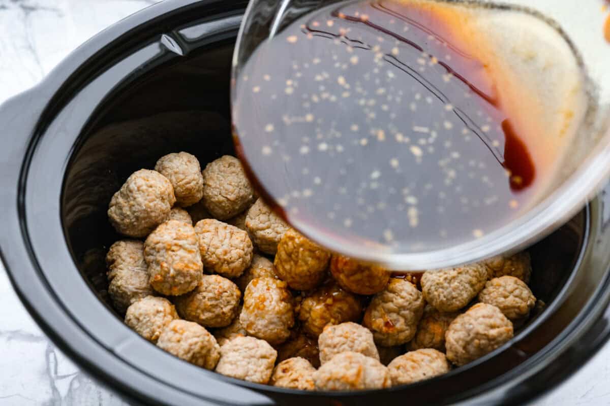 Slow cooker bourbon meatballs are an easy, set-it-and-forget-it favorite. Frozen meatballs simmer in a sweet, sticky bourbon sauce, making them perfect for an appetizer or a simple dinner with just 10 minutes of prep. Someone pouring the sauce over the top of the meatballs.