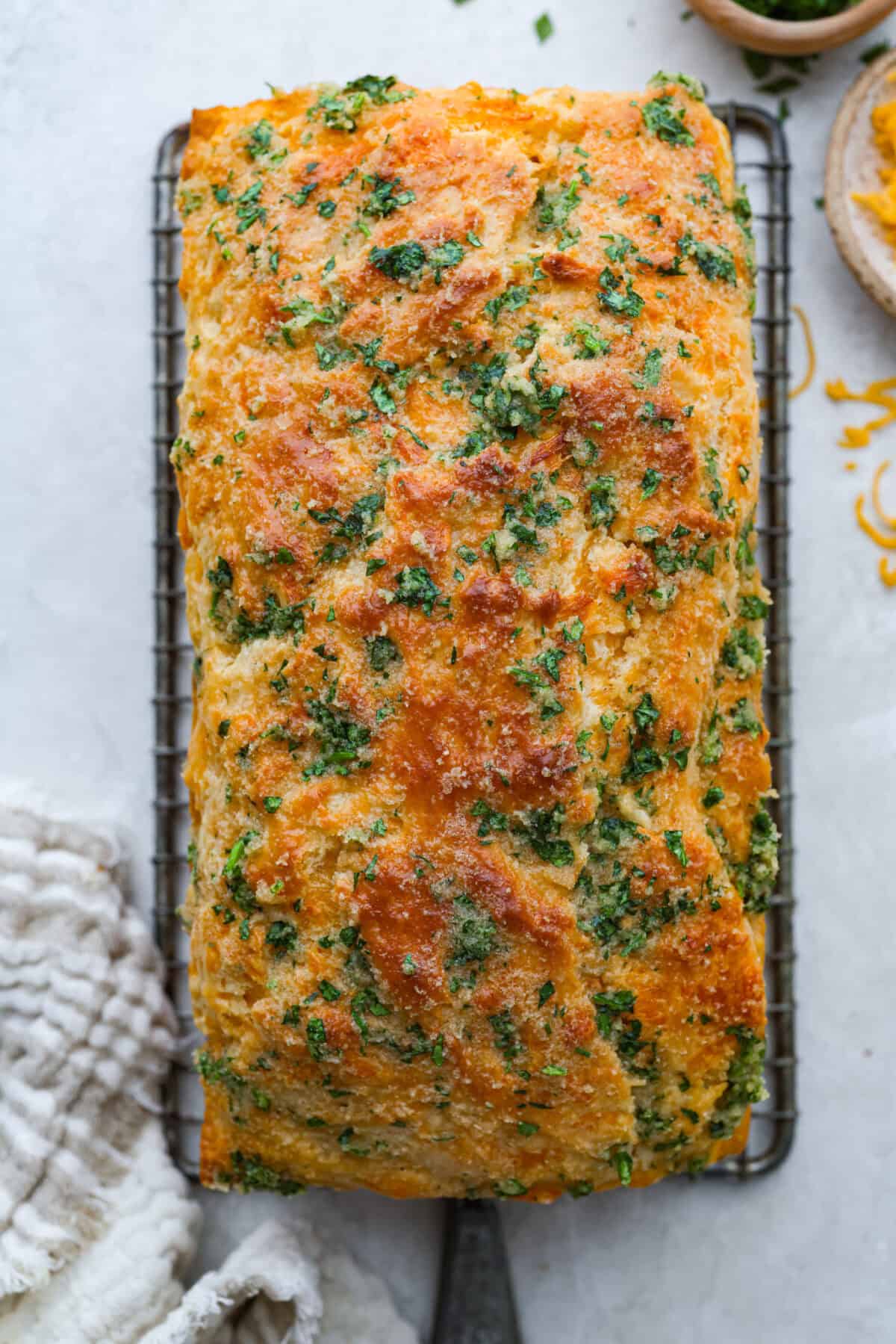 If you love my Red Lobster Biscuits, this Cheddar Bay Biscuit Bread has the same buttery, garlicky cheddar flavor, just mixed and baked right in a bread pan. Slice it warm and serve it with basically anything. It's a must-make! Trust me! Overhead shot of the cheddar bay biscuit bread loaf baked and cooking on a wire rack.