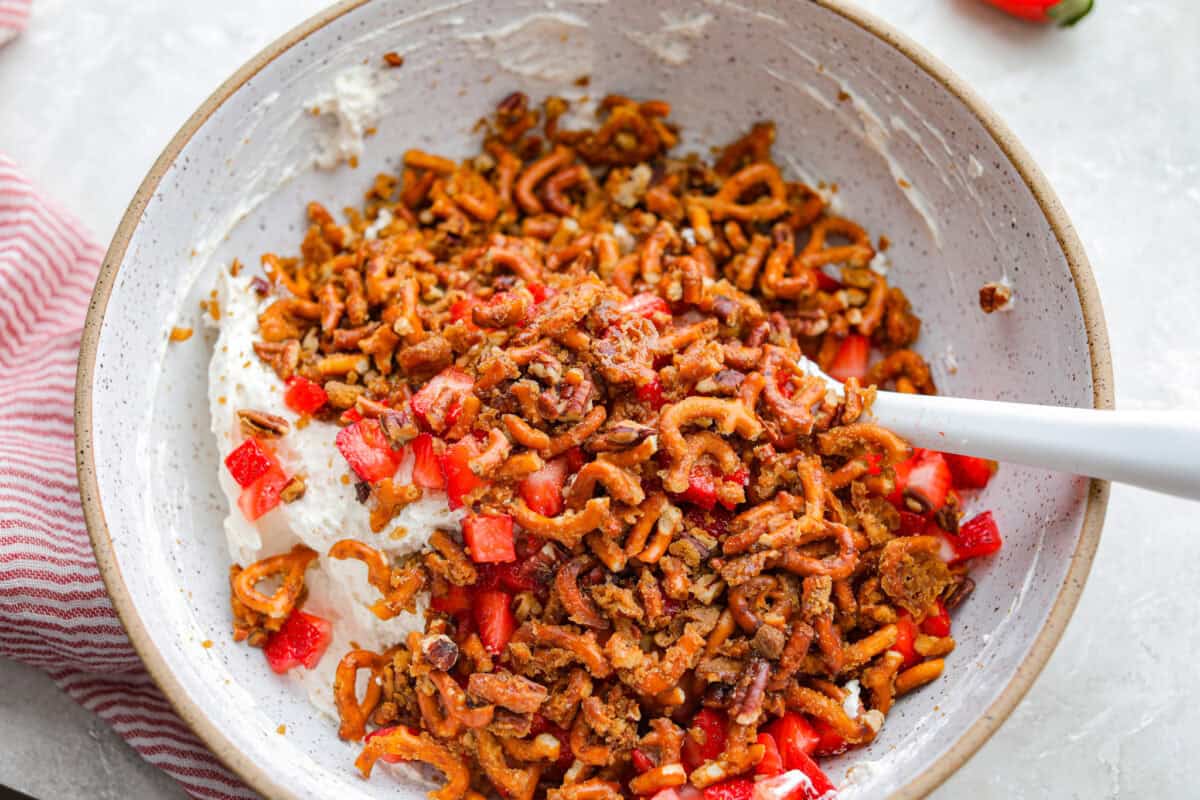 Crack pretzel salad is the shortcut version of my popular strawberry pretzel salad! It has all the sweet, salty, creamy goodness you love, but this version comes together in less than 15 minutes for an easy dessert everyone goes crazy for. All of the strawberry pieces, pretzel mixture, and cream cheese mixture added to a bowl to be stirred together.