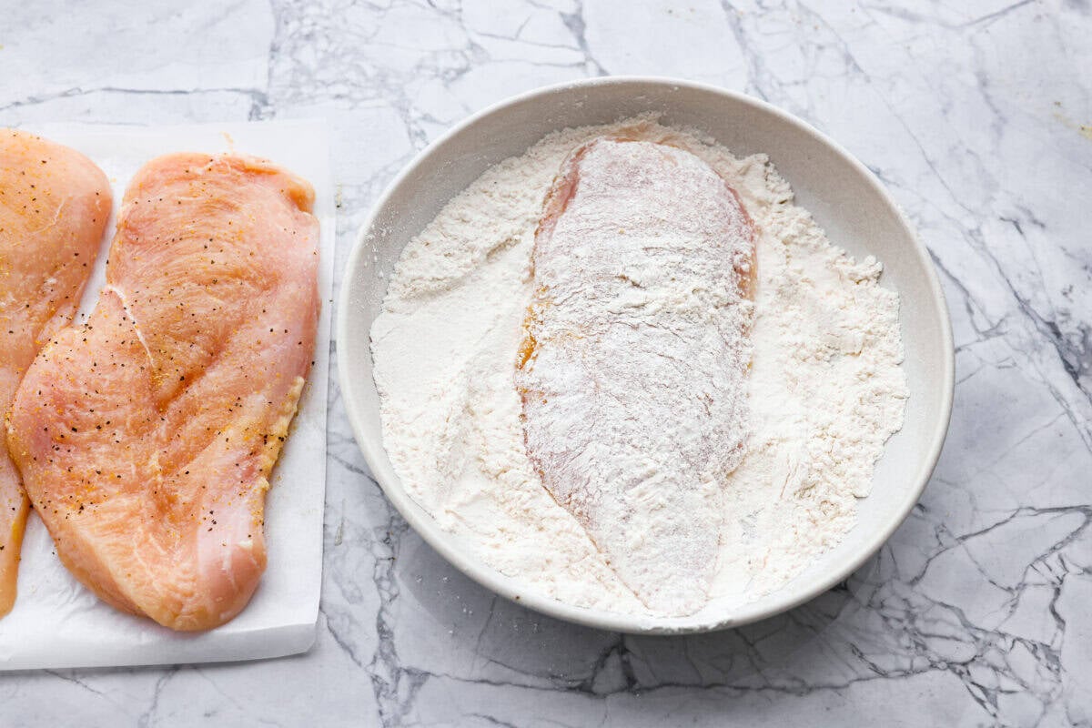 This lemon butter chicken is about to become your new weeknight secret weapon. One skillet, simple ingredients, and less than 30 minutes to the most golden, saucy, creamy chicken you've ever made. Chicken breast coated in flour in a dish.