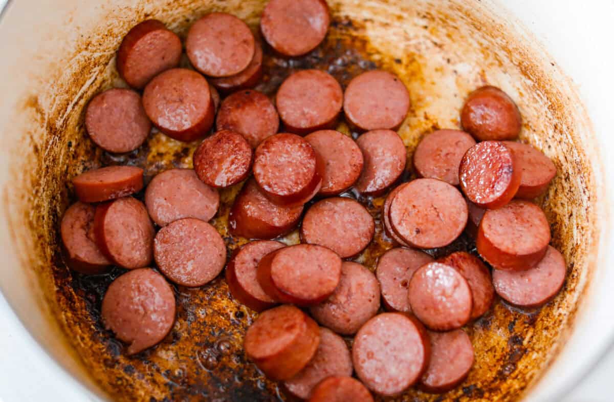 If you're bringing a dish to the cookout, make it these loaded baked beans. Three kinds of meat, two kinds of beans, and a sauce that is smoky, sweet, and full of depth. They disappear every single time. Sausage slices cooking in a large pot.