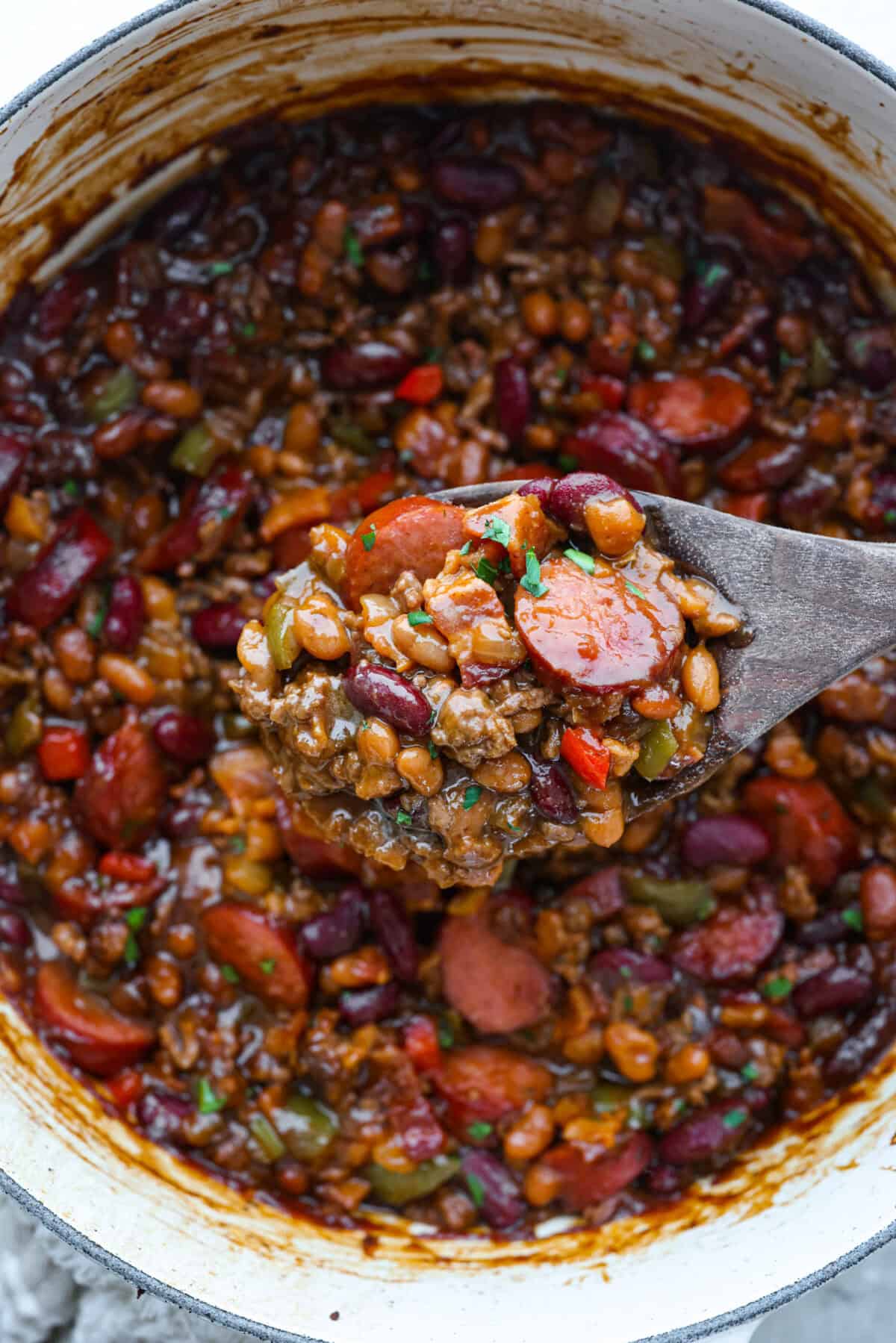 If you're bringing a dish to the cookout, make it these loaded baked beans. Three kinds of meat, two kinds of beans, and a sauce that is smoky, sweet, and full of depth. They disappear every single time. Pot of loaded baked beans with someone lifting a scoop out with a wooden spoon.