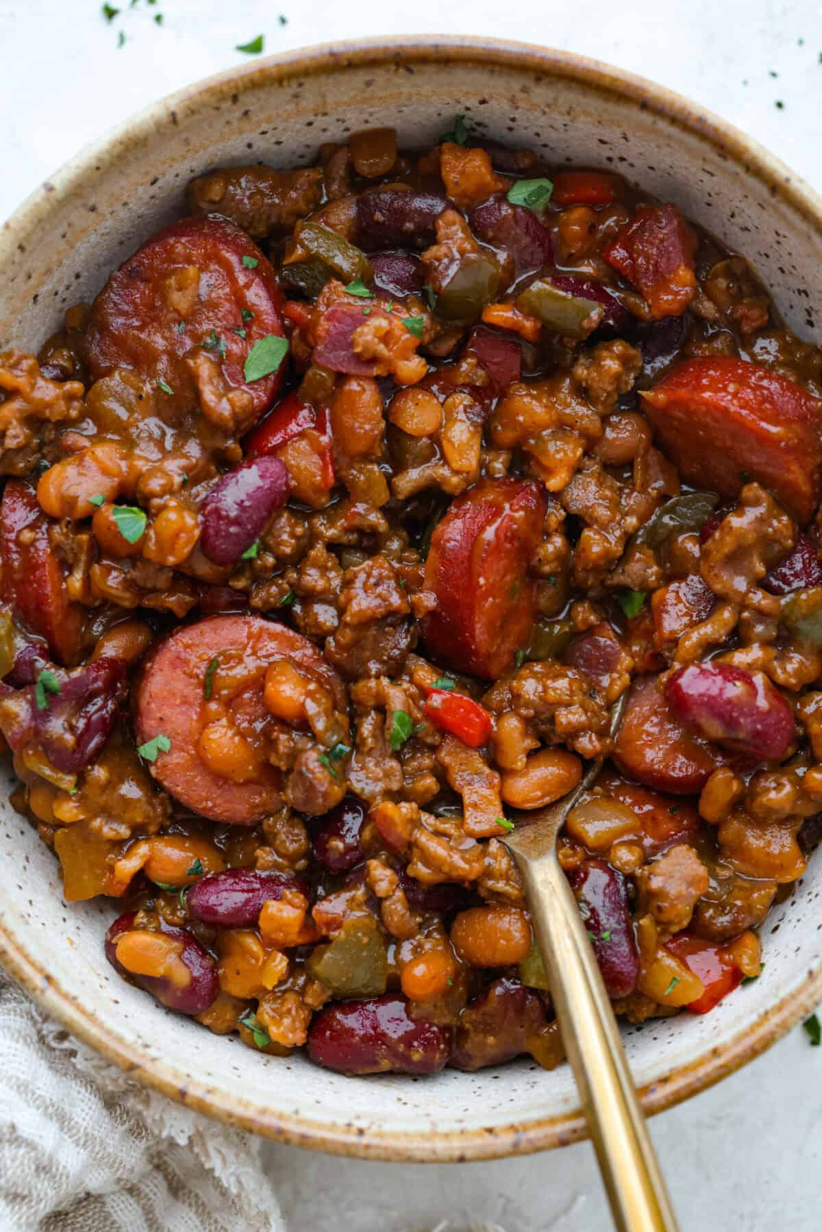 If you're bringing a dish to the cookout, make it these loaded baked beans. Three kinds of meat, two kinds of beans, and a sauce that is smoky, sweet, and full of depth. They disappear every single time. Bowl of loaded baked beans with a gold spoon.