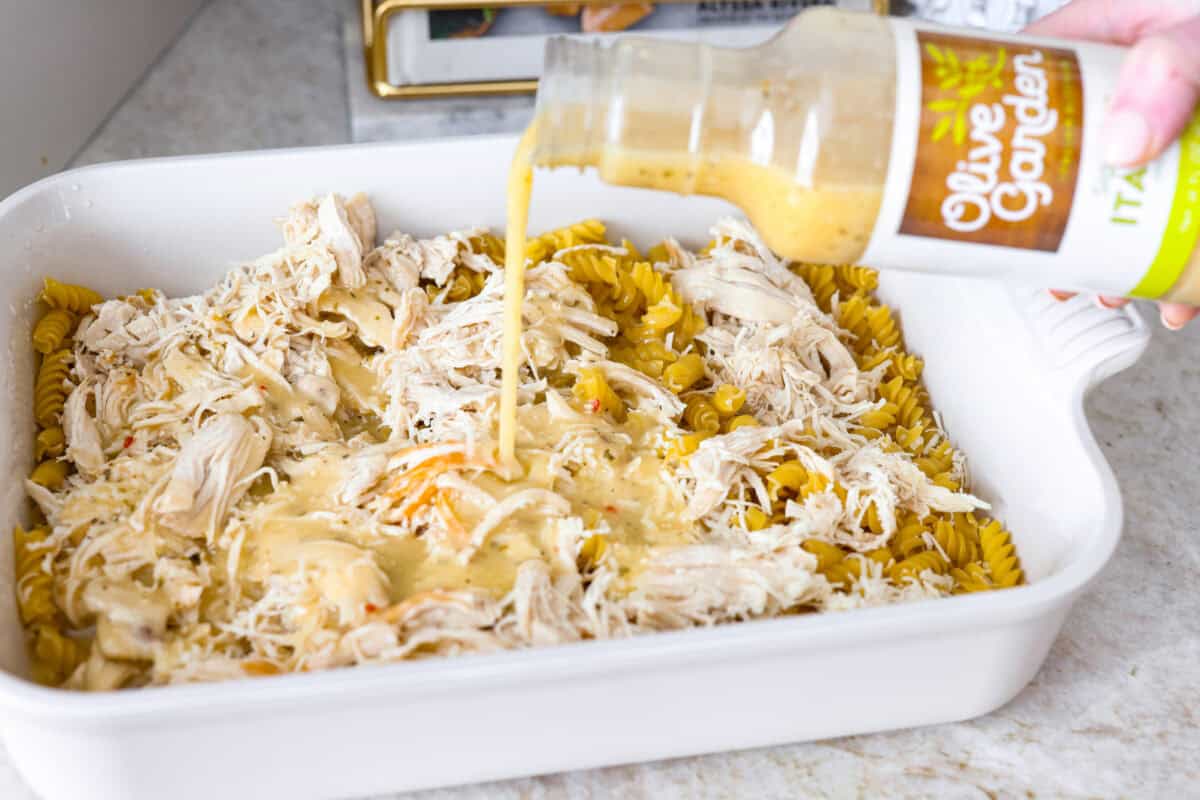This baked Olive Garden chicken is pure comfort food made easy! It uses Olive Garden Italian dressing and just 5 other ingredients to create a creamy, cheesy pasta bake everyone loves. Quick prep, one dish, and big flavor in every bite! Someone pouring Olive Garden dressing over the chicken and pasta.